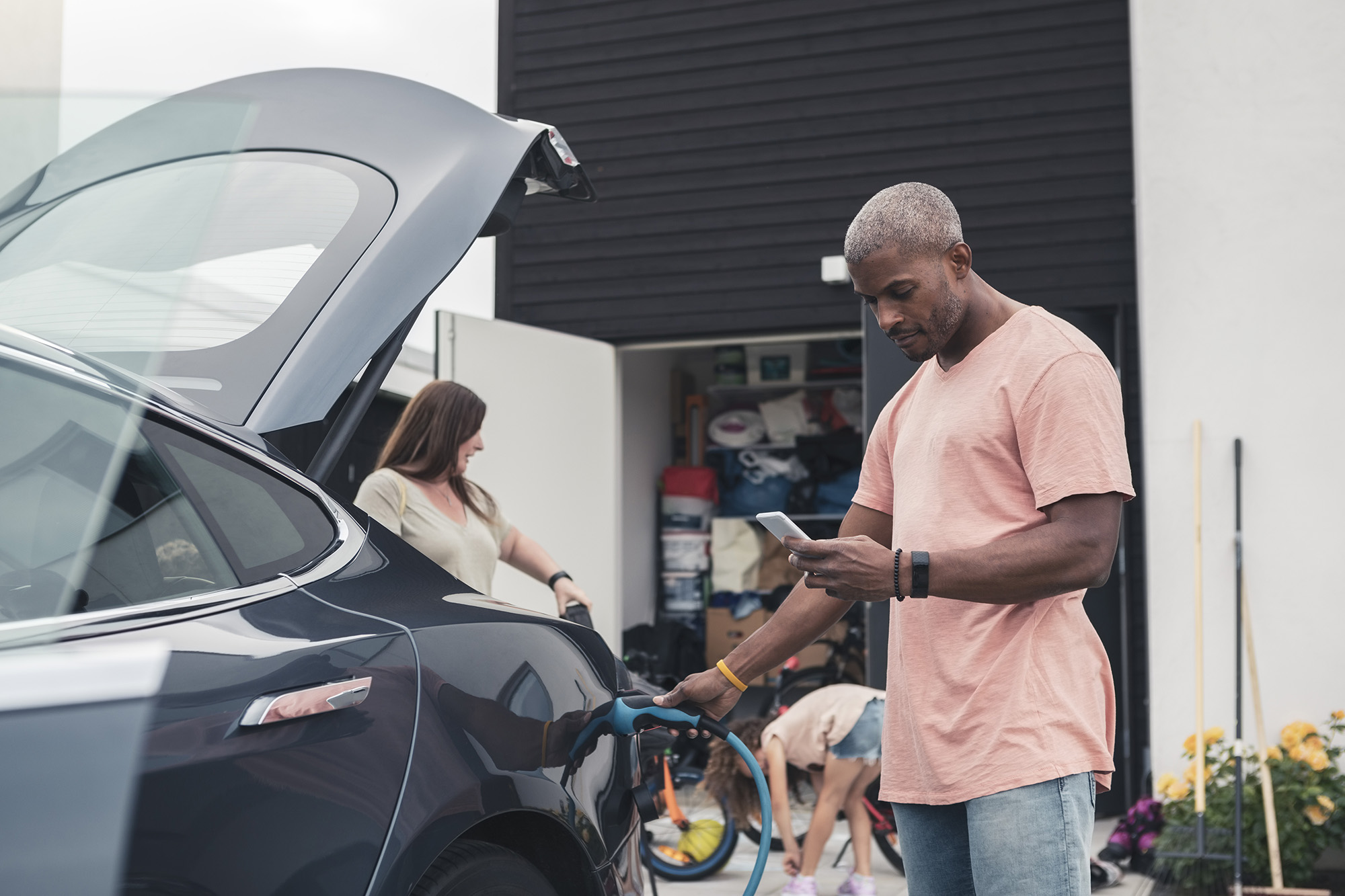 Man plugging in his EV looking at his cell phone while his family plays in the garage behind him