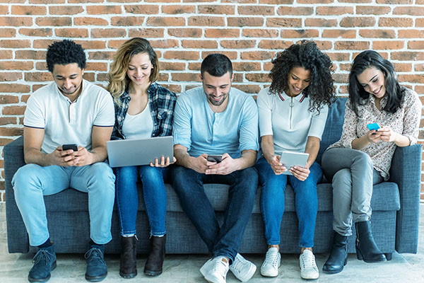 Five friends seating on a couch and looking at their laptops and mobile phones