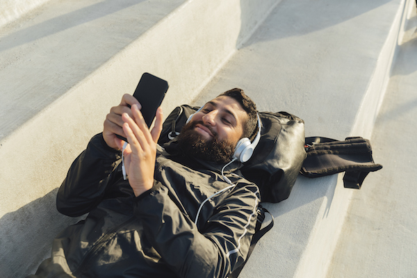 GettyImages-1127355140 Man lying on steps playing with smart phone and using headphones