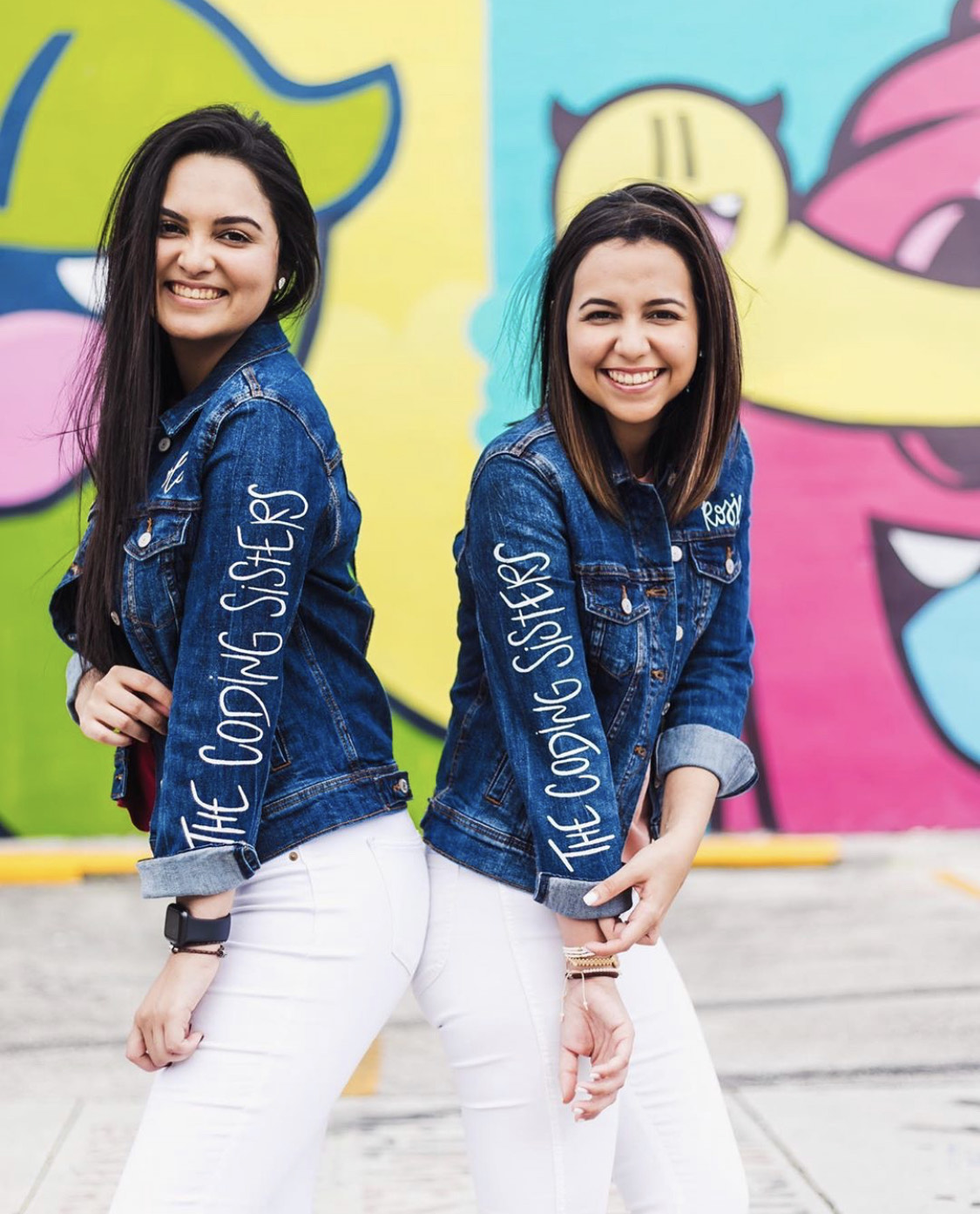 Sisters stand back to back in denim jackets smiling at camera