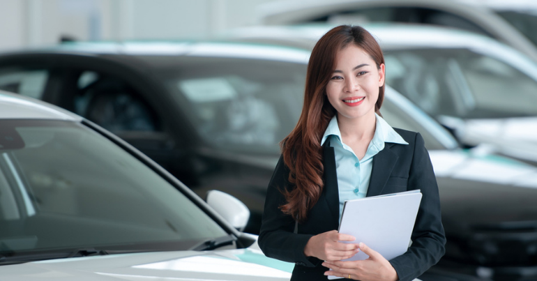 female car dealer with clipboard in hand standing in front of car