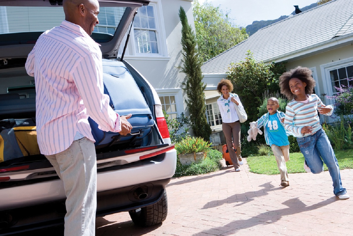 Family in Driveway Man unpacking the trunk of the car while his kids run towards him and wife walks with a suitcase
