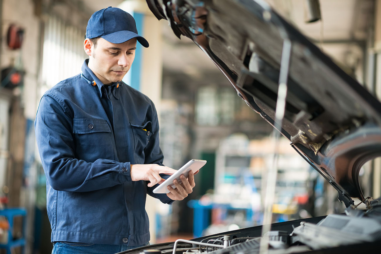 A mechanic dressed in blue checking the car engine and holding an ipad